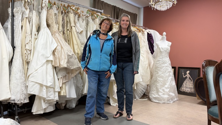 Twee vrouwen staan in een bruidswinkel omringd door trouwjurken, met een paspop en een kroonluchter op de achtergrond.