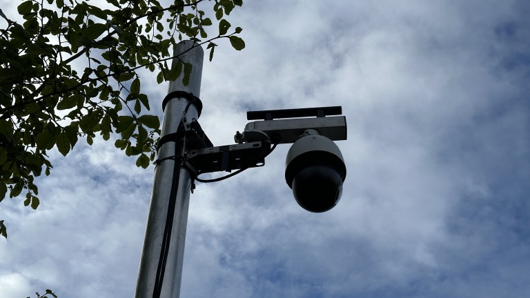 Bewakingscamera bevestigd aan een paal naast een boom, gefotografeerd tegen een bewolkte hemel.