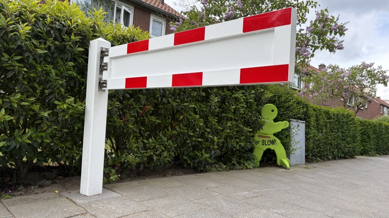 Wit-rode slagboom langs een trottoir met op de achtergrond een groene pop met "SLOW!" in een groene haag.