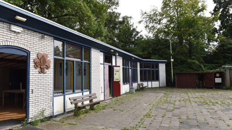 Bakstenen gebouw met blauwe kozijnen en houten bankjes, omgeven door groene bomen, met een houten scoutingembleem naast de ingang.