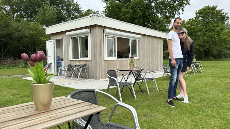 Een koppel staat samen voor een houten gebouw met een terras, waarin tulpen in potten op tafels staan, omgeven door groene bomen.