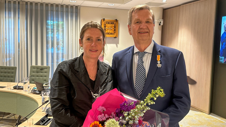 Een man en een vrouw poseren in een vergaderruimte, de man draagt een koninklijke onderscheiding en de vrouw houdt een boeket bloemen vast.