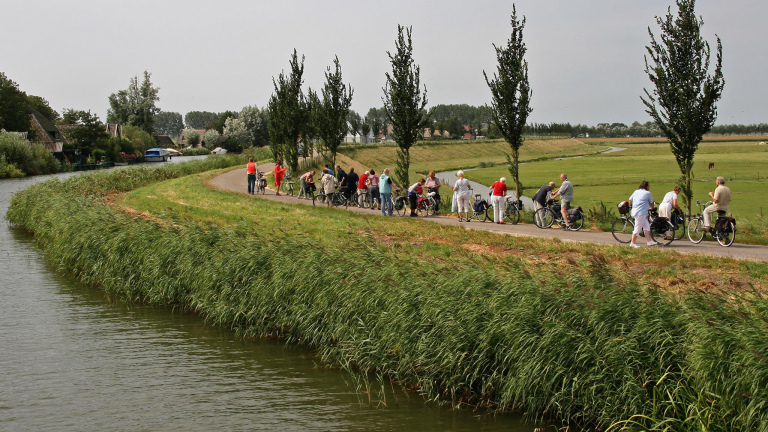 Groep mensen fietst langs een kanaal in een groen, landelijk landschap met enkele bomen.
