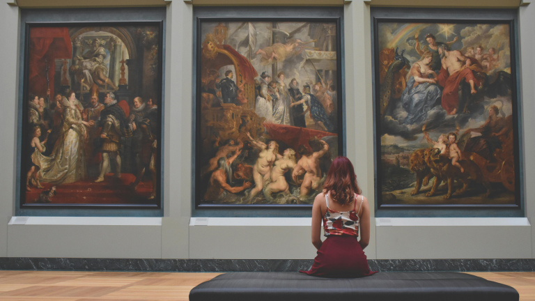 Een vrouw zit op een bank in een kunstgalerie en bewondert drie grote schilderijen met historische en mythologische scènes.