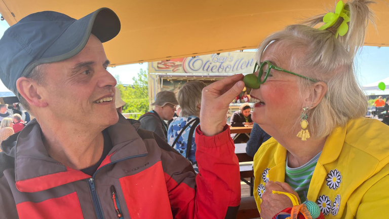 Twee mensen zitten buiten te lachen, terwijl de vrouw een groene clownsneus en bril draagt. Op de achtergrond een oliebollenkraam.