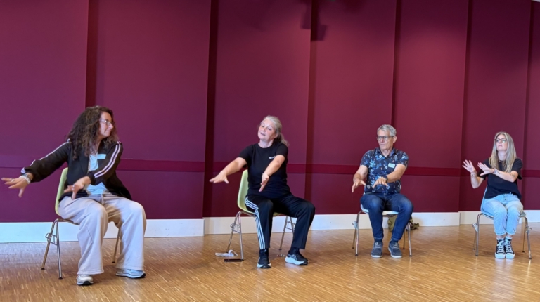 Een groep mensen zit in een kring op stoelen en voert gezamenlijke armbewegingen uit in een zaal met rode muren.