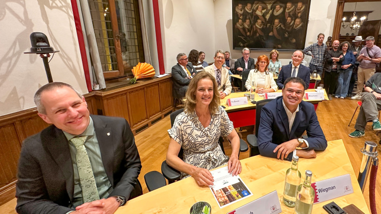 Mensen zitten aan een tafel in een formele setting, met naamkaartjes en glazen water voor zich, omringd door andere aanwezigen.