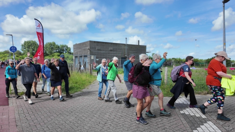Een groep mensen wandelt over een stoep langs een fietspad, sommige zwaaien naar de camera op een zonnige dag.