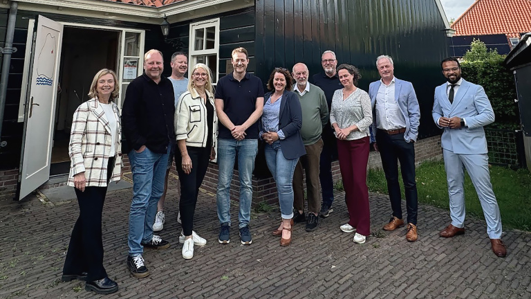 Een groep van elf mensen poseert buiten voor een gebouw, deels onder een overkapping, lachend in informele kleding.