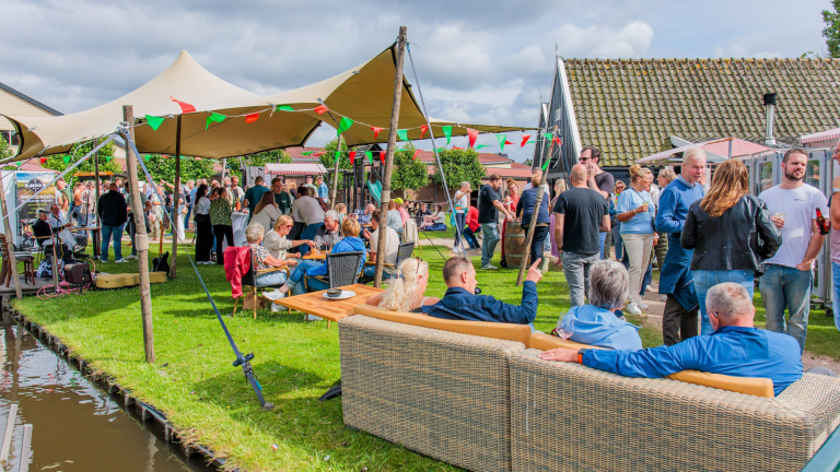 Mensen genieten van een buitenfeest met tenten, zittend op banken en stoelen, onder slingers van kleurrijke vlaggetjes.