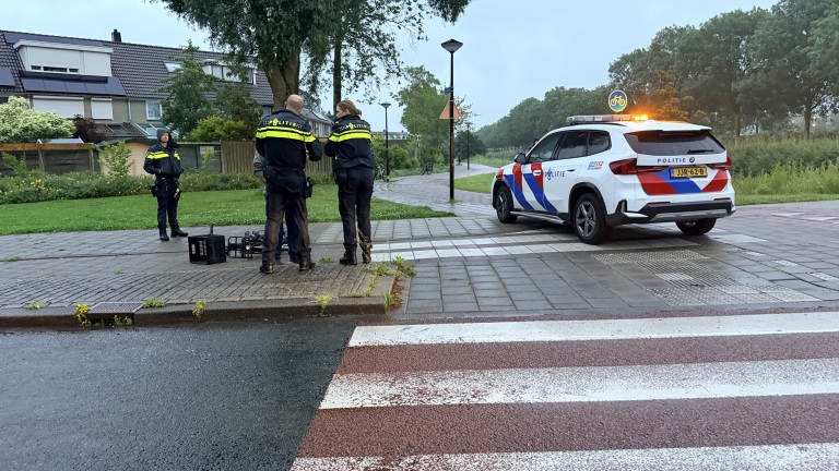 Drie politieagenten staan op een stoep naast een politieauto met zwaailichten aan bij een kruispunt.