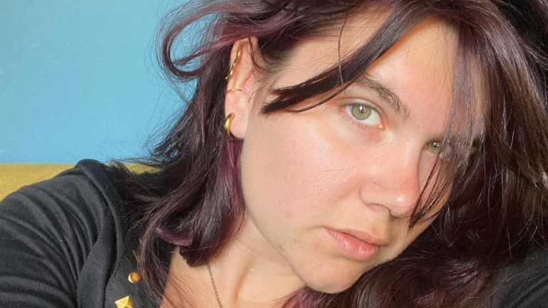 Persoon met donker haar en gouden oorringen tegen een blauwe achtergrond.
