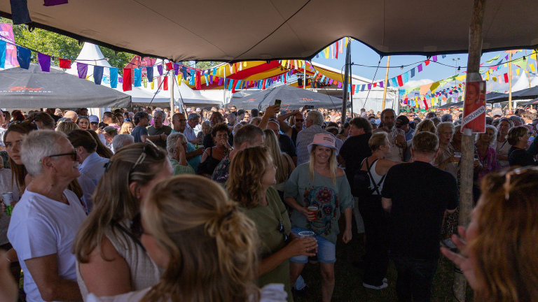 Feestelijke menigte onder een tent met kleurrijke vlaggen, mensen genieten van het evenement en het zonnige weer.