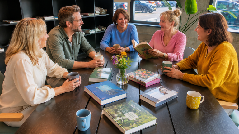 Vijf mensen zitten rondom een tafel met boeken en drinken koffie, terwijl ze samen praten en lachen.