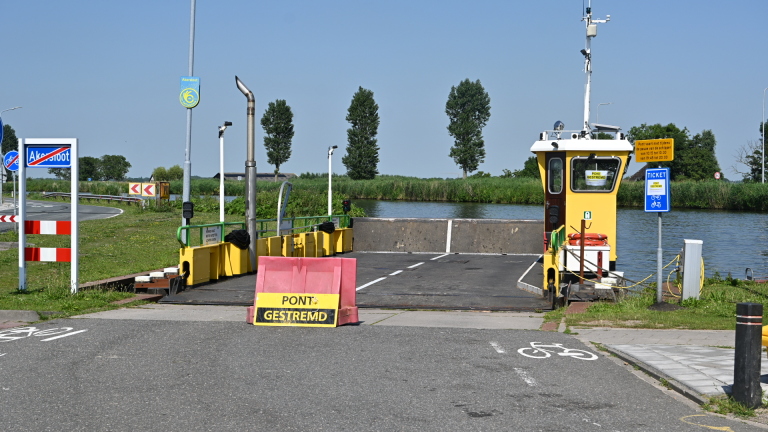 Geel veerbootje genaamd Akersloot met auto aan boord, vastgemaakt aan een pier, op de achtergrond enkele huizen.