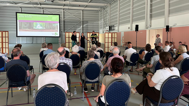 Mensen zitten op stoelen in een sporthal en kijken naar een presentatie op een groot scherm.