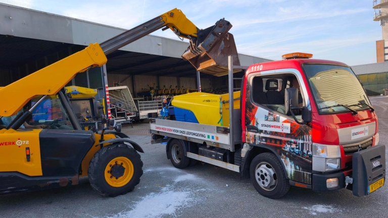 Een gele bulldozer laadt een geel-blauwe machine op de laadbak van een rode vrachtwagen met "Stadswerk 072" op de zijkant.