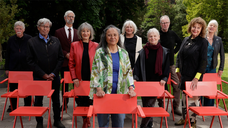 Een groep oudere mensen poseert buiten, achter felrode stoelen, omringd door veel groen.