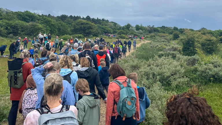 Een grote groep mensen wandelt over een zandpad door een groen landschap onder een bewolkte hemel.