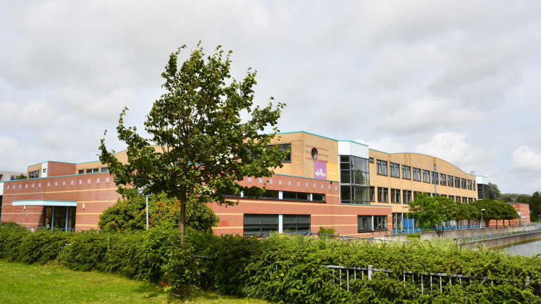 Een modern gebouw van baksteen en glas, omgeven door een groen landschap met een boom op de voorgrond en een gracht aan de zijkant, onder een bewolkte hemel.