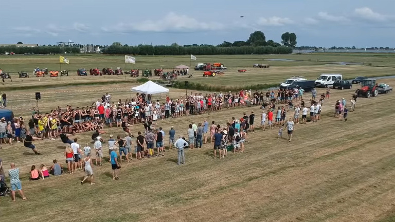 Grote groep toeschouwers op een grasveld bij een touwtrekwedstrijd, met tractoren en tentjes op de achtergrond