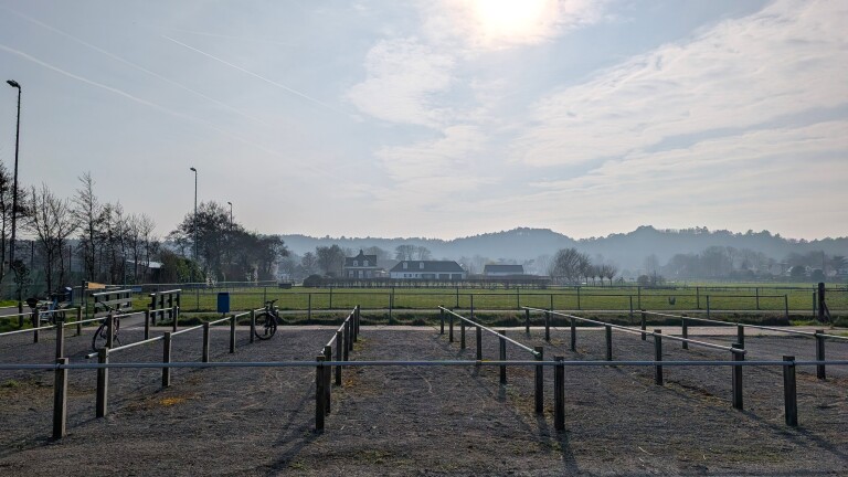 Leeg fietsenrek op een open ruimte met enkele fietsen en een landschap van velden en huizen onder een bewolkte hemel.