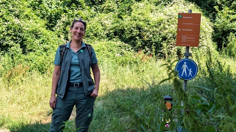 Vrouw in groene wandelkleding met rugzak naast bord Waarderhout met tekst Vrij wandelen, in een groen bosgebied