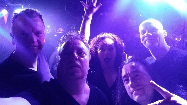 Selfie van vijf mensen in een club met paars-blauw licht; één steekt een hand omhoog en één maakt een peace-teken