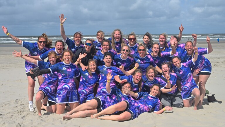 Juichend team in paarse sterrenhemel-shirts poseert op het strand, met zee en wolkenlucht op de achtergrond