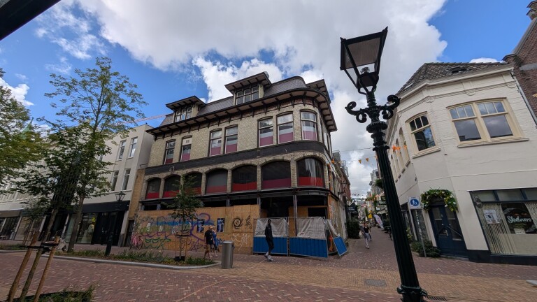 Straatbeeld met historisch pand in renovatie, afgeschermd met houten platen en bouwhekken, onder blauwe lucht met lantaarnpaal