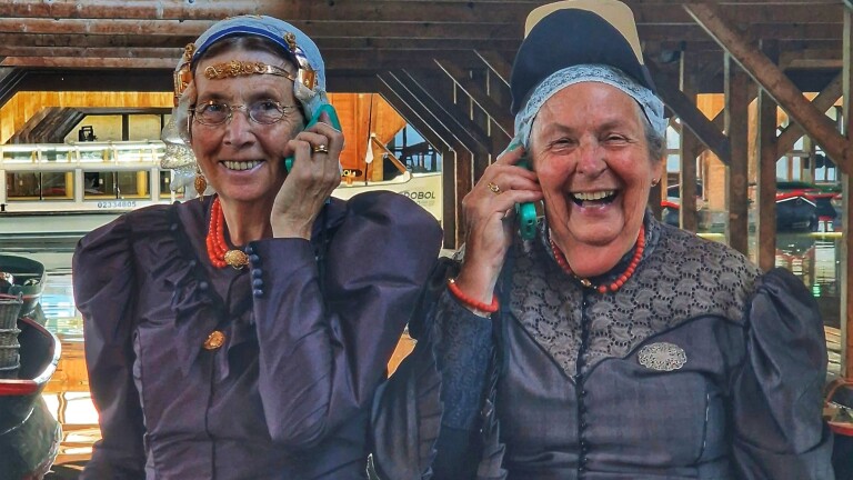 Twee lachende oudere vrouwen in historische klederdracht bellen met een groene telefoon, met boten op de achtergrond