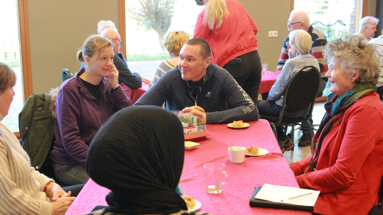 Groep mensen praat en lacht aan een tafel met roze kleed, met koffie en gebak, in een buurthuisachtige ruimte
