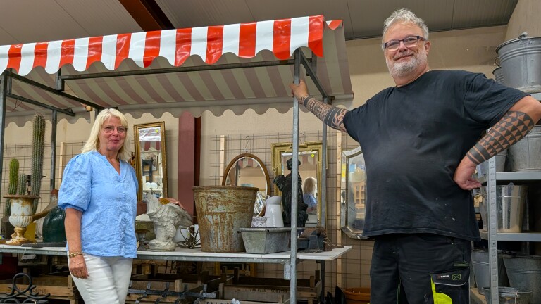 Twee mensen poseren bij een marktkraam met rood-wit gestreepte luifel, omringd door spiegels, potten en decoratie