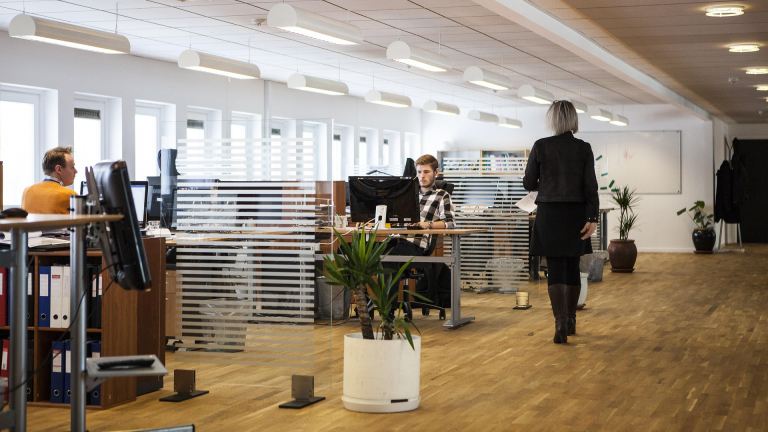 Open kantoor met werknemers achter computers, glazen wanden en een vrouw die door de ruimte loopt
