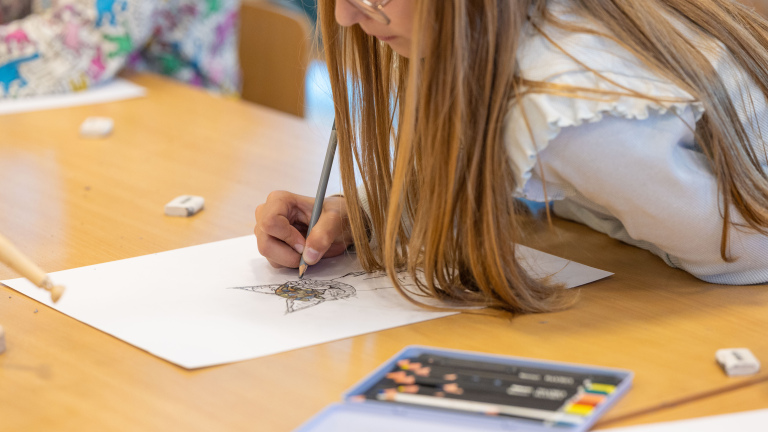 Meisje met lang haar tekent met potlood op papier aan een tafel, met gum en kleurpotloden op de voorgrond