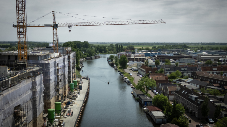 Een bouwplaats aan een kanaal met hijskranen en steigers; woningen en groene bomen langs het water.