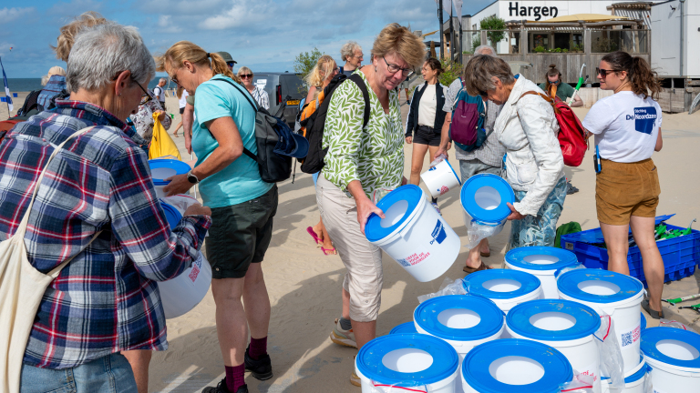 Vrijwilligers op het strand verzamelen witte emmers met blauwe deksels voor een opruimactie van Stichting De Noordzee
