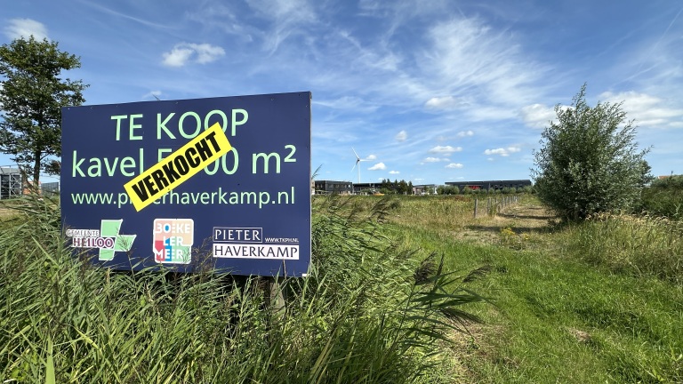 Bord met “Te koop” en sticker “Verkocht” bij een grasveld, met gebouwen en een windmolen op de achtergrond
