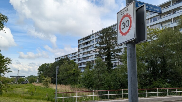 Verkeersbord met snelheidslimiet van 30 km/u voor een appartementencomplex, met op de achtergrond een grotendeels verborgen molen.