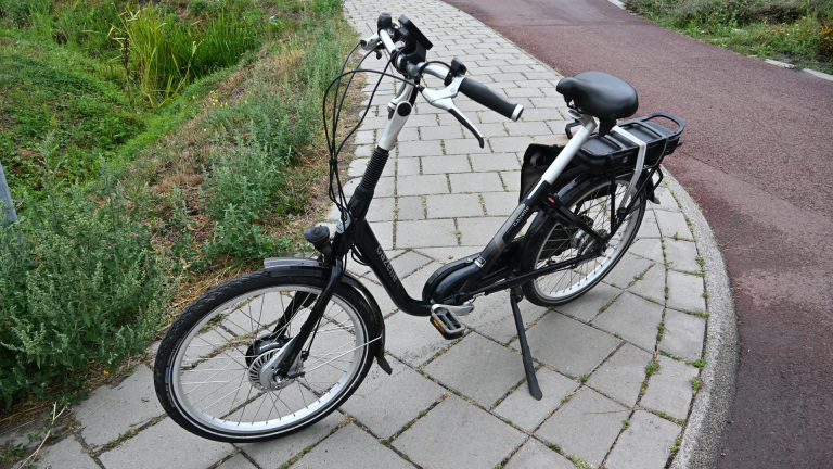 Zwarte elektrische stadsfiets met lage instap en accu op de bagagedrager, geparkeerd op een stoep langs een fietspad