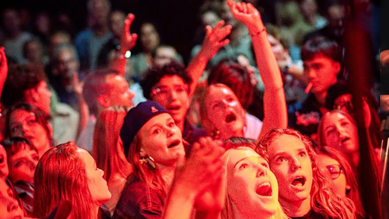 Jong publiek bij een concert onder rood licht, met opgeheven handen en zingende, juichende gezichten