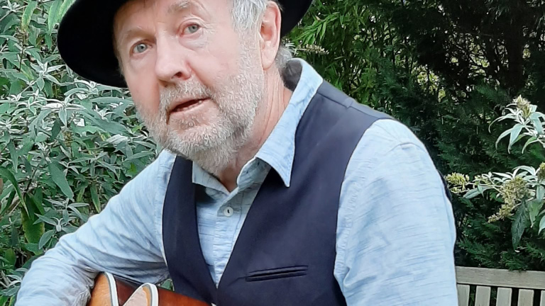 Oudere man met grijze baard en zwarte hoed speelt gitaar op een bankje tussen groen struikgewas
