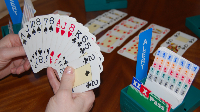 Hand met speelkaarten en bridge-biedbox op tafel, met uitgelegde kaarten op de achtergrond