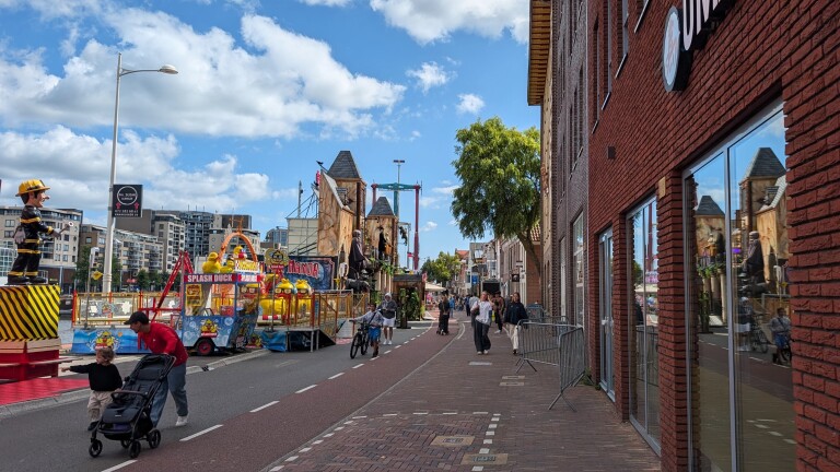 Straat met kermisattracties zoals Splash Duck en Manor, met wandelaars en een kind op een fiets naast bakstenen panden