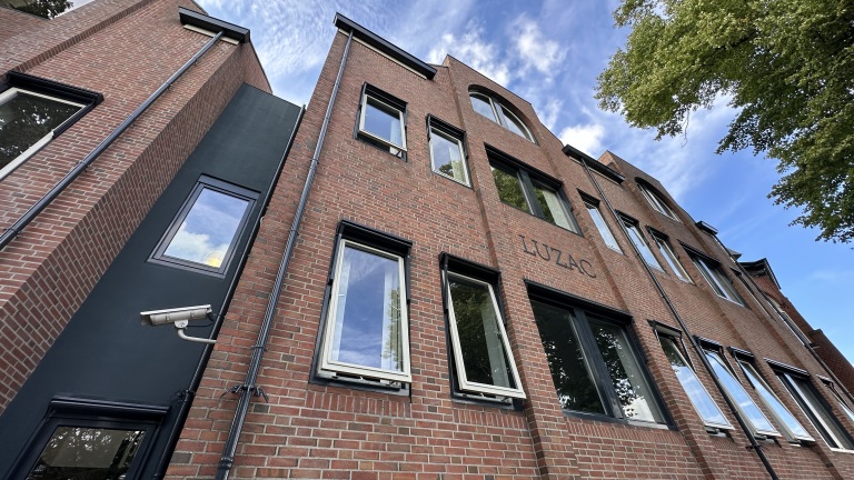 Rood bakstenen gebouw met torentje en windvaan naast glazen aanbouw, met grote boom en passerende voetgangers