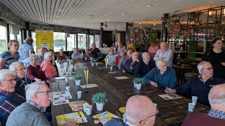 Een grote groep oudere mensen zit aan een lange tafel in een gemeenschappelijke ruimte, omringd door decoratieve planten.