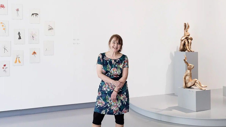 Vrouw in bloemige jurk in een galerie, met tekeningen aan de muur en twee gouden beeldjes op sokkels