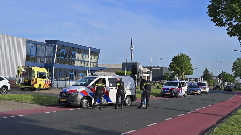 Politieagenten bij meerdere politievoertuigen en een ambulance naast een modern gebouw langs de weg.