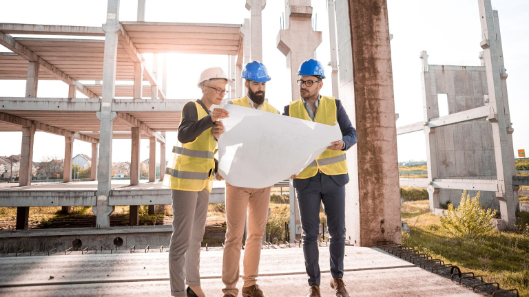 Drie bouwprofessionals met helmen en veiligheidsvesten bekijken een bouwtekening op een constructieterrein
