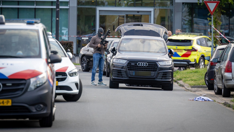 Gemaskerde agent met vuurwapen bij zwarte auto met open achterklep, met politie- en ambulancevoertuigen op straat